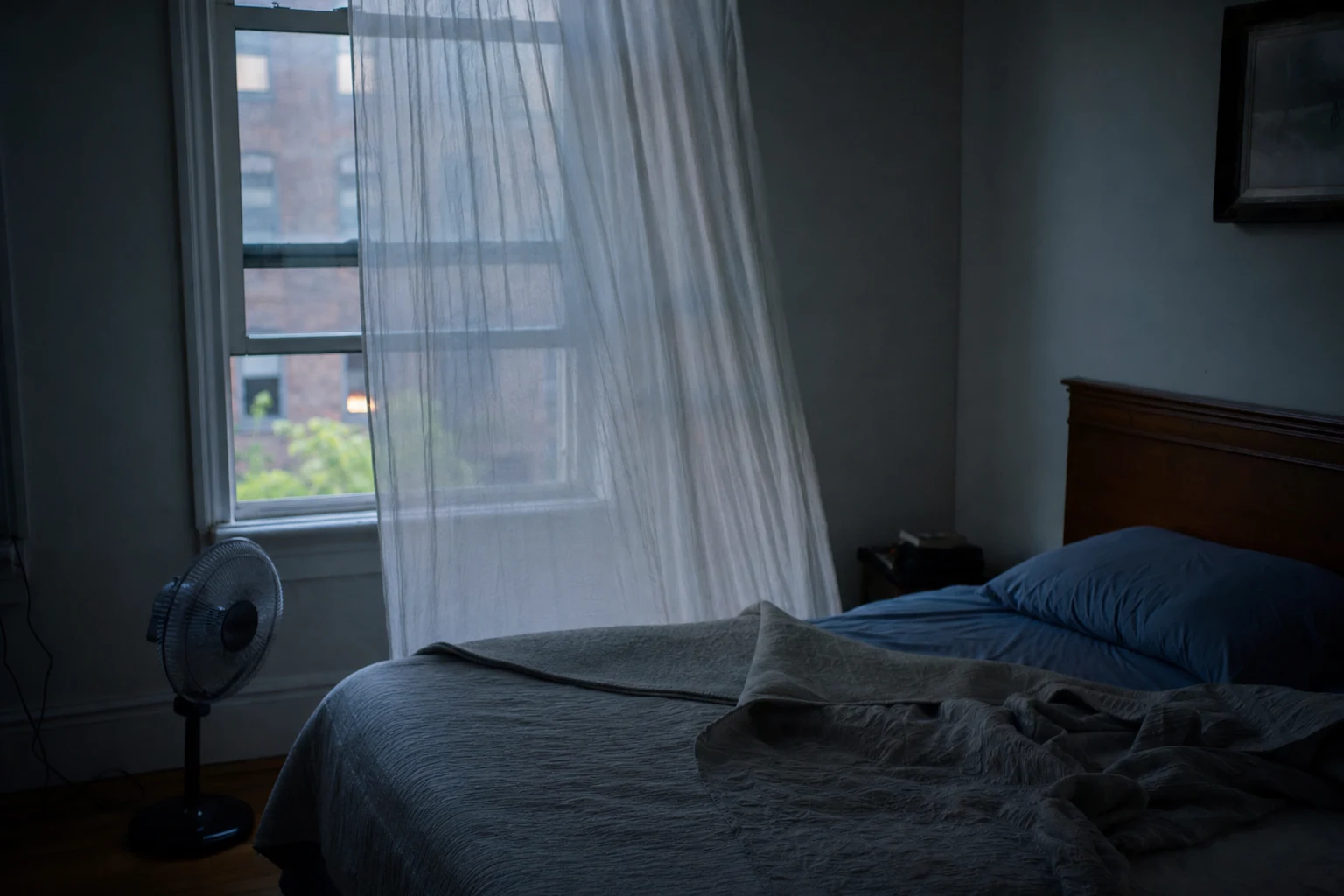 A cool, dim bedroom with the window open a crack and a light blanket folded back