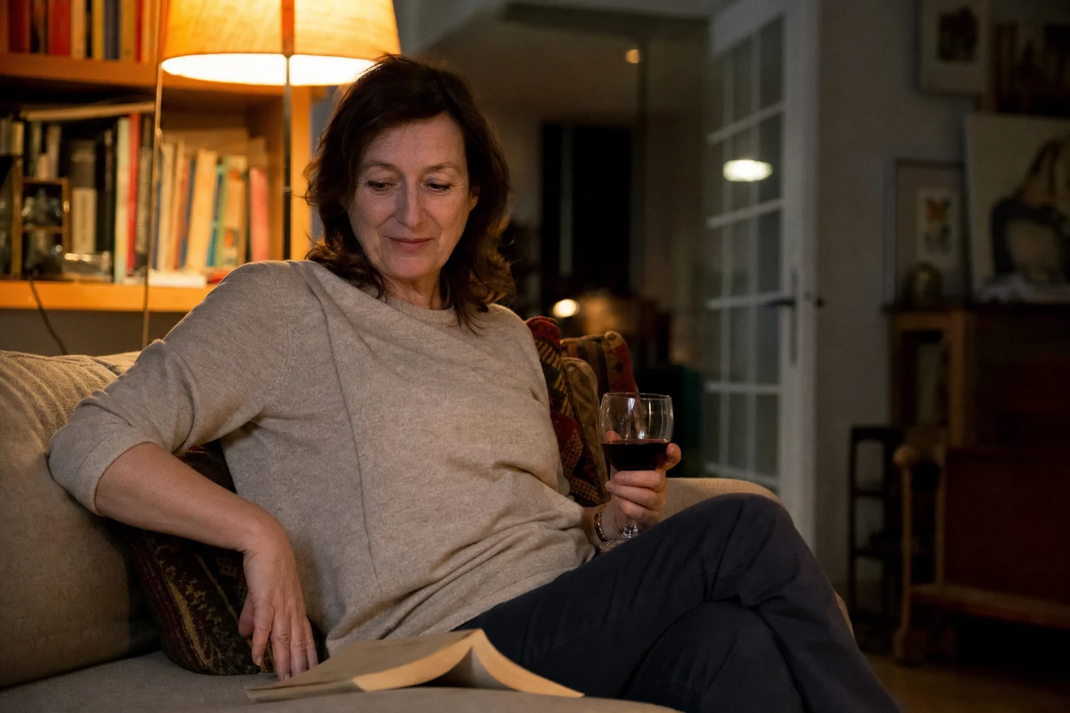A woman in her fifties holding a glass of red wine on the sofa in the evening, a book open beside her