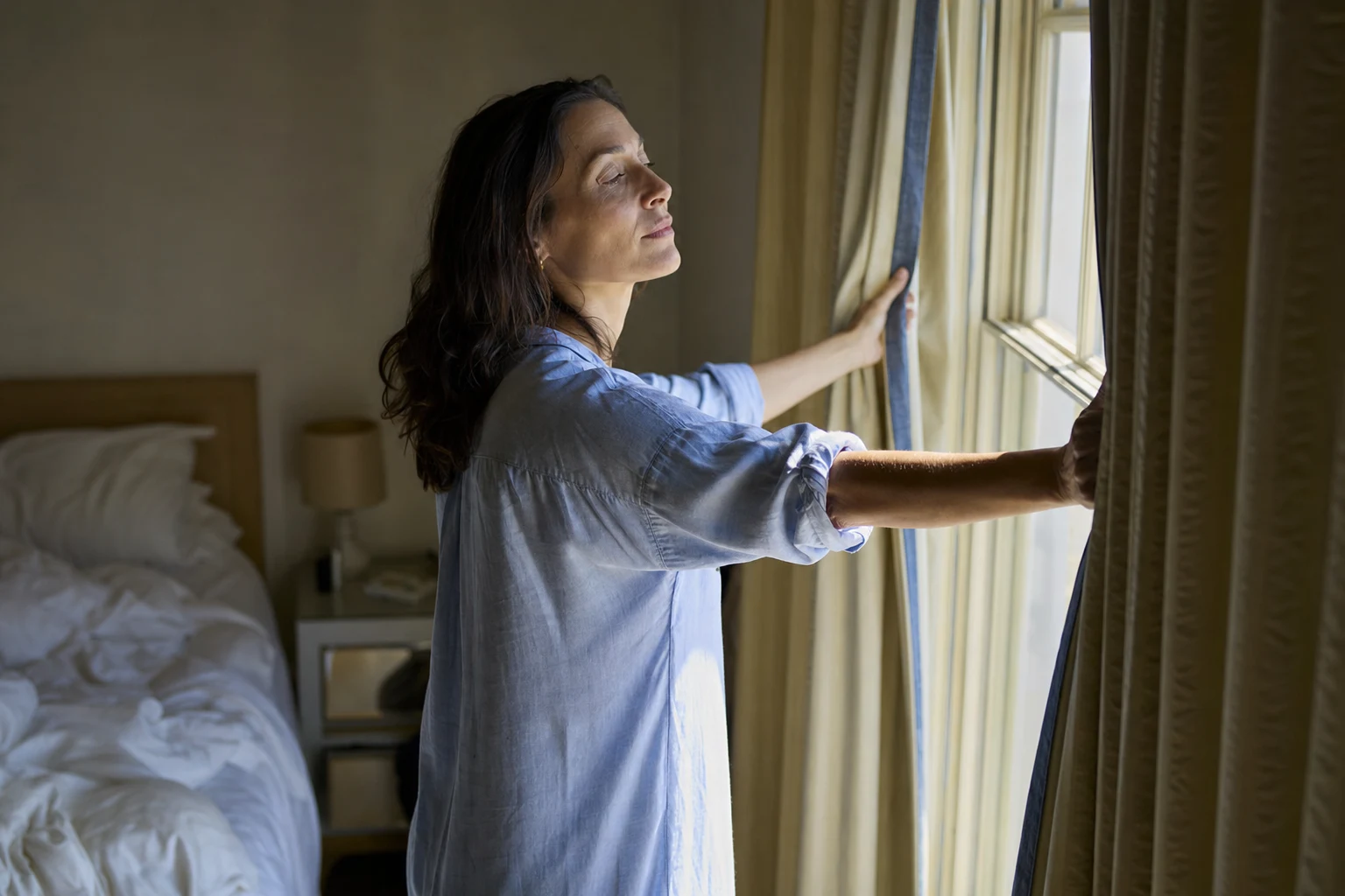 A woman in her forties opening the bedroom curtains to morning light